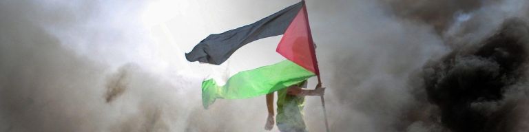 Man carrying a Palestinian flag surrounded by fire and smoke.