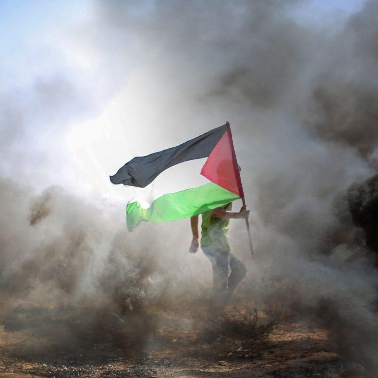Man carrying a Palestinian flag surrounded by fire and smoke.